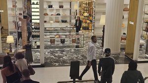 A sales advisor stands alone behind a display counter in a luxury department store featuring Celine branding, marble floors and gold shelving, while customers walk through the foreground in motion.